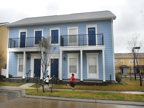 Photograph of a two-story duplex, with a porch and second floor balcony attached to each unit. Two children running along a sidewalk surrounded by grass are visible in the foreground.