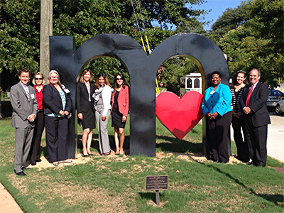 Image of nine individuals gathering around an approximately 8 foot tall lower case letter “m”, containing a heart in the second curve.