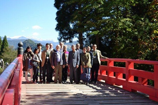 Image of 13 individuals standing on a bridge with red rails; the mountains of Tateyama are visible in the background.