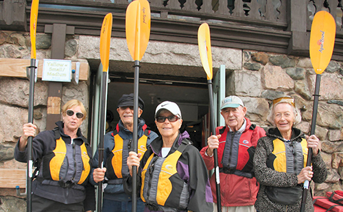 A group of five men and women who are members of Newton at Home ready to go kayaking, prepared with water resistant clothing, life vests, and paddles.