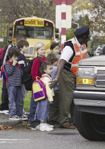 A group of young school children being helped to cross a busy street by a school crossing guard.