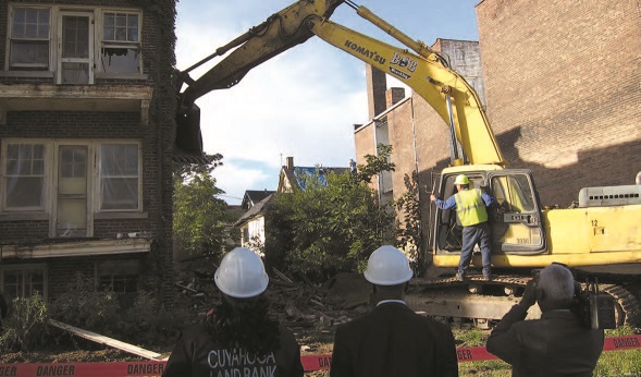 A work crew in hardhats operating heavy equipment to demolish a vacant, abandoned brick apartment building.