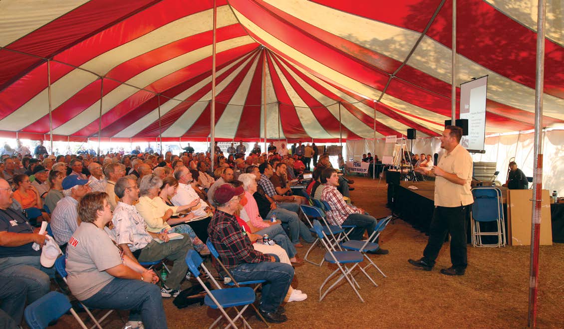 A group of people seated on folding chairs in a tent, listening to a speaker.