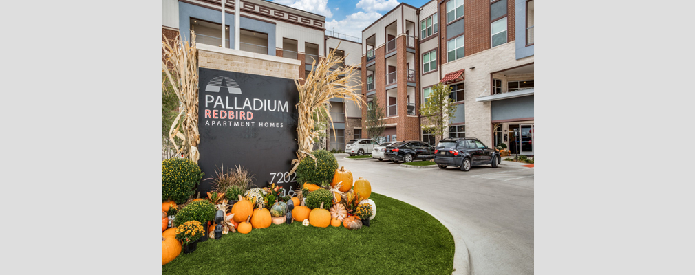 An entrance sign with pumpkins and shrubs at its base with a full parking lot and a four-story building in the background.