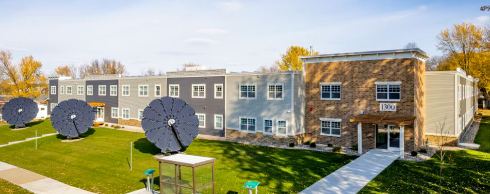 Front view of a two-story multifamily development on a grassy lot. Three ground-mounted solar panel arrays and a bus stop are in front of the building.