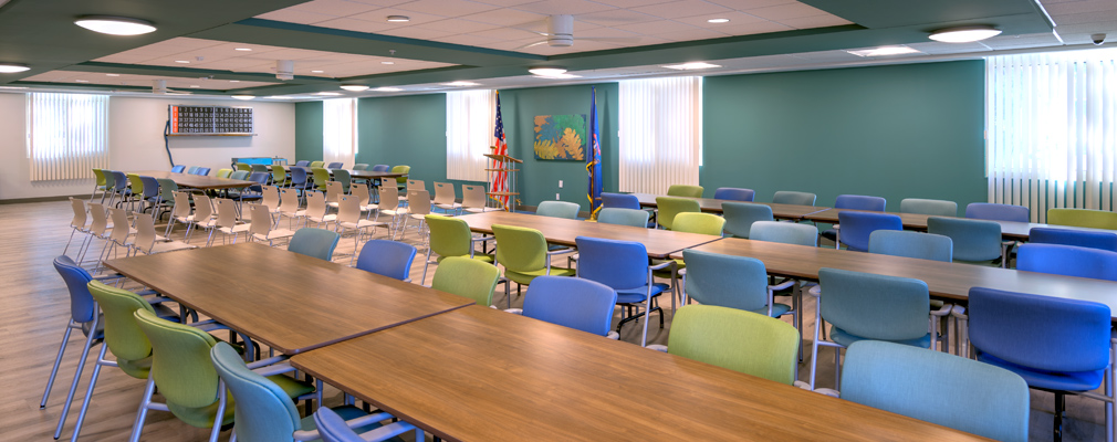 Room with three rows of chairs and desks with two flags in the background, windows, and curtains.