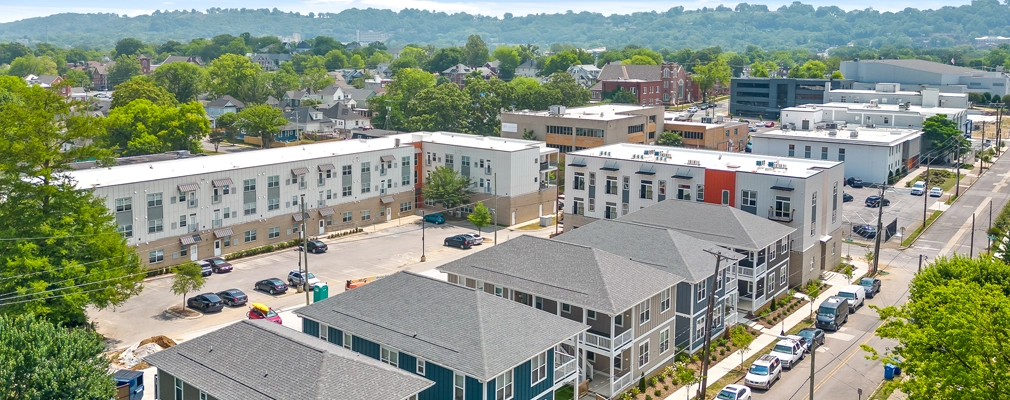 A low aerial photograph of a row of 5 two-story quadplexes with pitched roofs and front porches, fronting a residential street with parked cars. Several three-story apartment buildings are in the background.