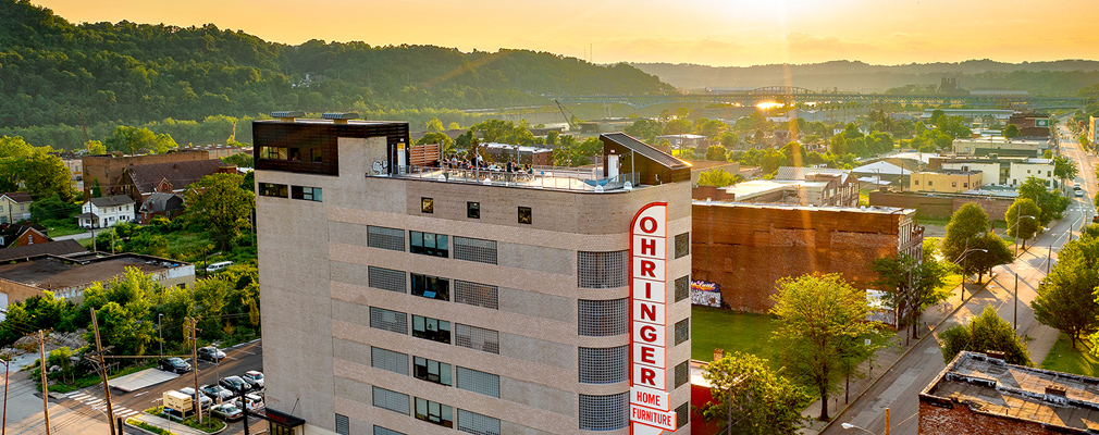 Low-angle aerial photograph of an eight-story building with a five-story blade sign that reads "Ohringer Home Furniture" in the foreground and the Monongahela River and mountains in the background.