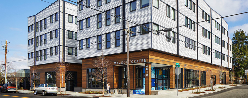 A photograph of a four-story building with a street, sidewalk, parked cars, and street trees in the foreground.
