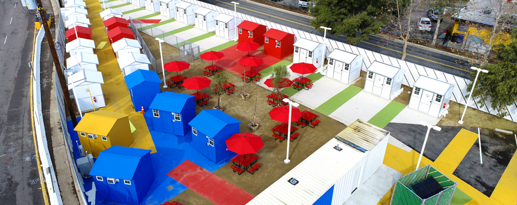  An aerial photograph of a brightly painted tiny home village surrounded by a white chain-link fence and white-painted highway barriers. 