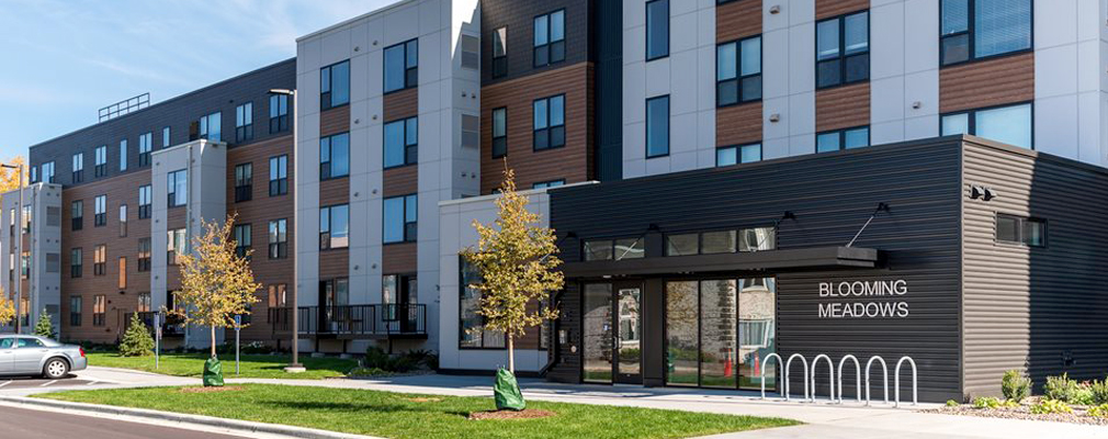 Front façade of a four-story apartment building with the sign "Blooming Meadows" on the wall.