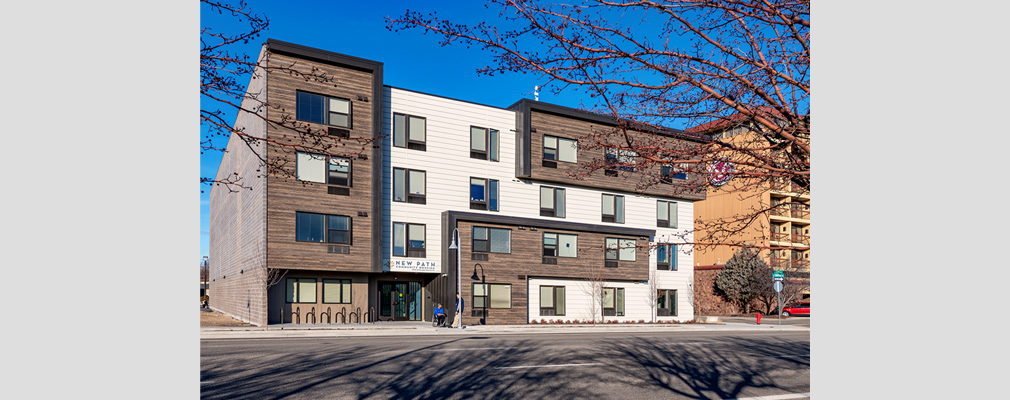 Photograph of the front façade of a four-story multifamily building, with a sign above the entrance reading, “New Path Community Housing.” 