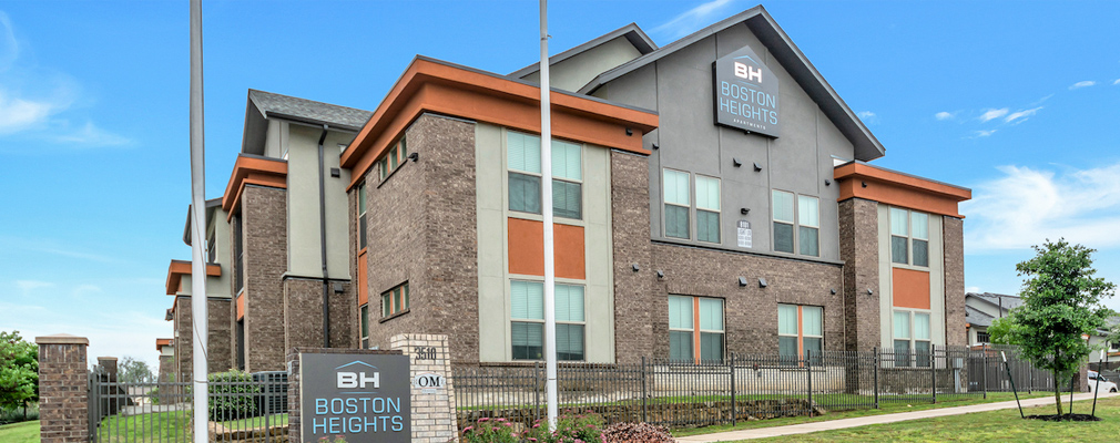 The street façade of a two-story apartment building, with a wall sign in the gable of the building and a monument sign in the foreground reading “BH Boston Heights.”