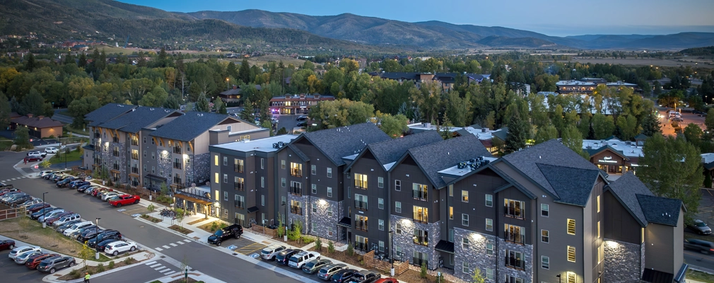 An aerial view of a four-story building surrounded by parked cars in front, a playground in the foreground, and mountains in the background.