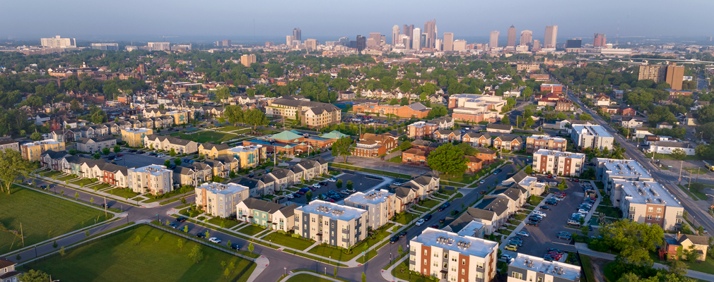 Aerial photograph of a mixed-use community covering several city blocks with apartment buildings, townhomes, and parking lots, with a downtown skyline in the background.