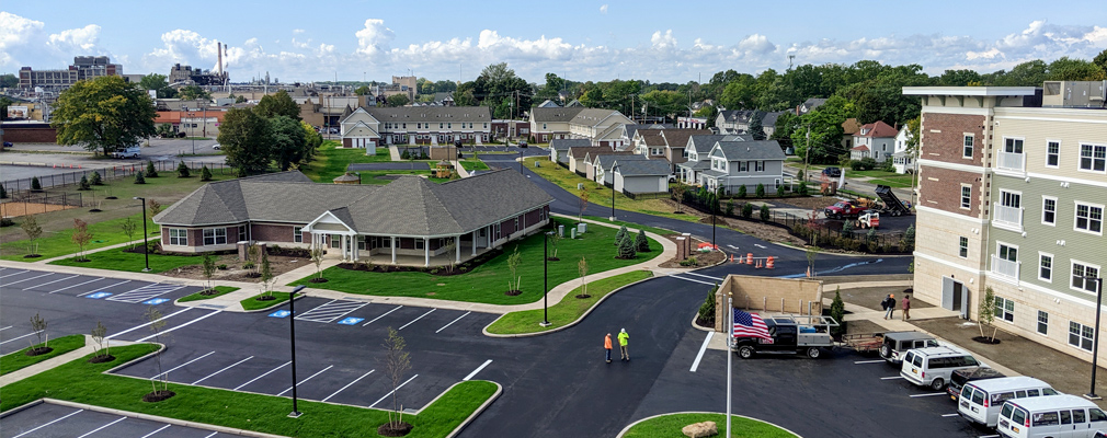 Photograph of a single-story clubhouse building (center left) and a portion of a four-story multifamily building (right), with single-family detached houses and townhouses in the background and surface parking in the foreground.