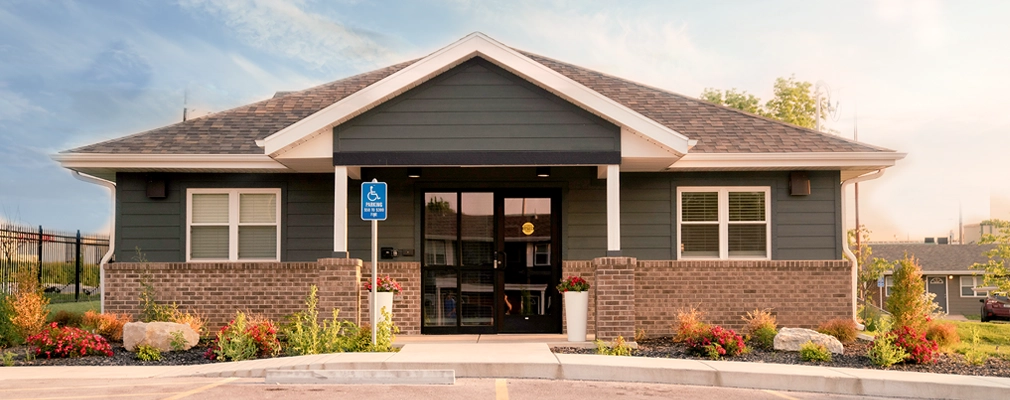A photograph of a single-story building with a gabled roof and a covered entrance flanked by landscaped planting beds, with parking spaces in the foreground.