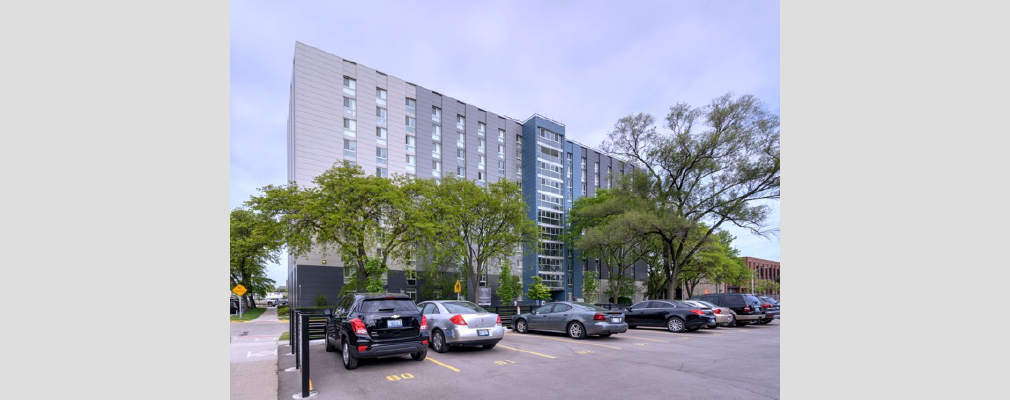 Photograph of an 11-story building lined with trees and facing a parking lot filled with cars.