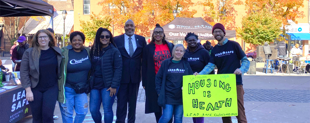 Eight smiling people posing in front of a Lead Safe Resource Center outreach booth, one holding a sign that reads ”Housing Is Health.”