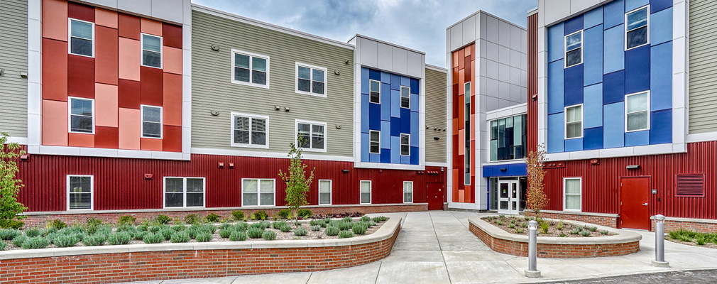 Photograph of the entrance to three-story multifamily building, with a landscaped courtyard in the foreground.