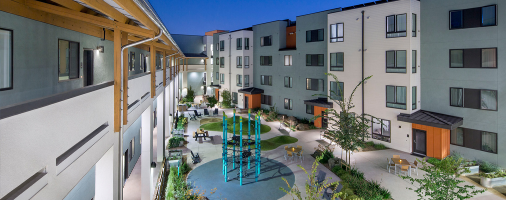 Photograph of a landscaped courtyard with play equipment and sitting areas framed by a four-story apartment building taken at dusk from an upper story.