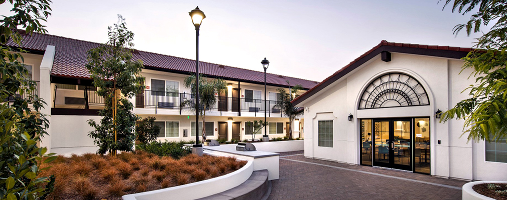 Photograph of a single-story, tile-roofed building with landscaping and grills in the foreground and a two-story walk-up apartment in the background.