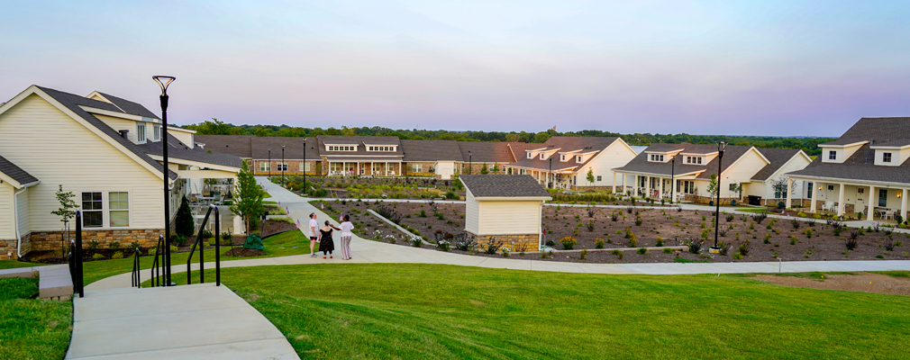 Several one-story residences framing a farming plot, with paved walkways and an expansive lawn in the foreground.