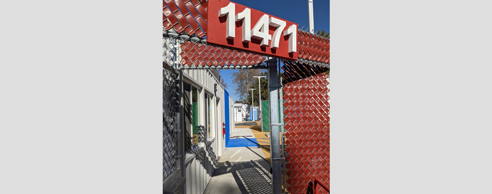  Photograph of a corrugated metal trailer, taken through an opening in a red chain-link fence.