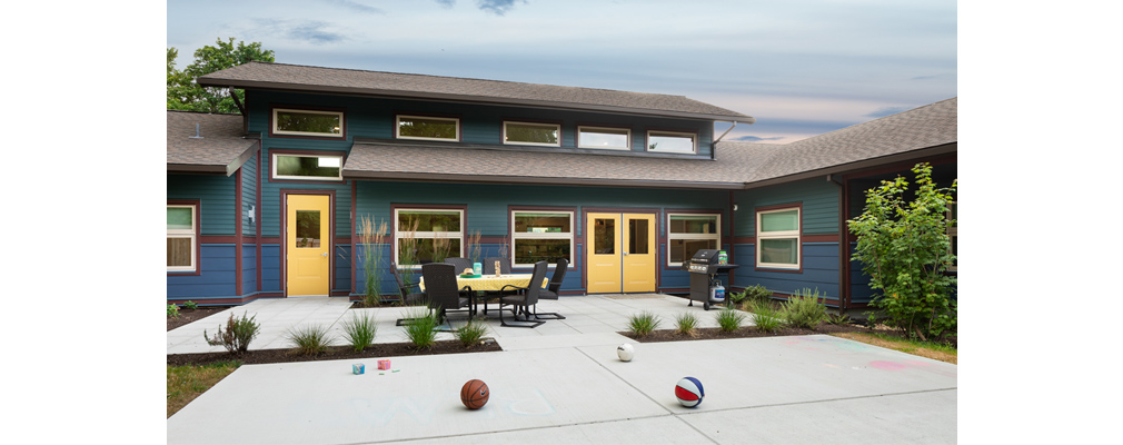 Rear façade of a one-story building standing behind a table and chairs set on a patio and sports balls lying on a court.