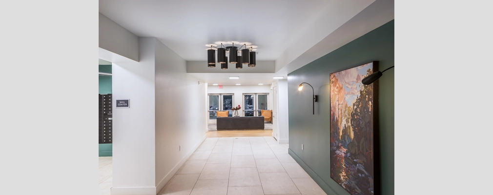 A hallway leading to a seating area with a sofa and chairs, with a painting and light fixture on the wall.
