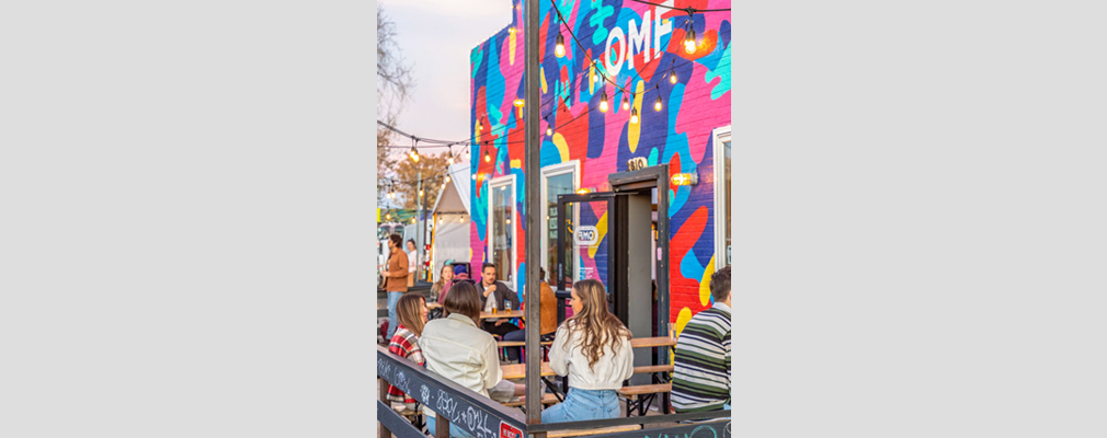 Photograph of people dining at tables in front of a restaurant with a mural on its entrance wall.  