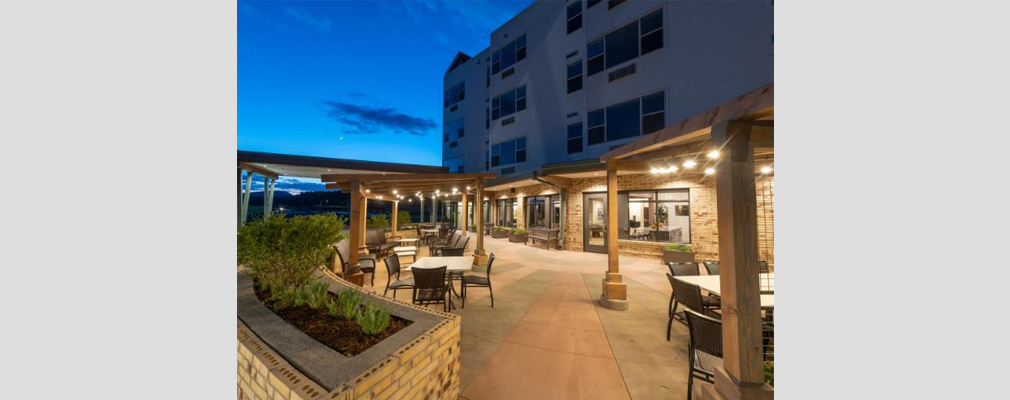 An evening view of a four-story building with a large stone patio containing chairs and tables underneath multiple pergolas.