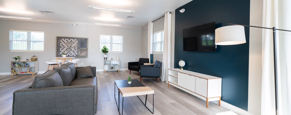 A photograph of a community room with a mounted television, sofa, lounge chairs, and coffee table in foreground and bookshelves and a table with chairs near windows in the background.