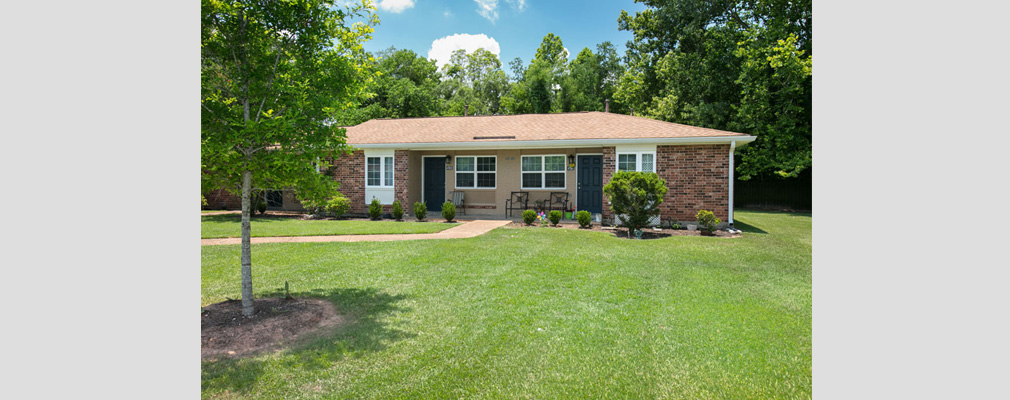 View of the front entrances of two units at Tranquil Estates, with a tree in the foreground and a walkway leading to a front porch with chairs.