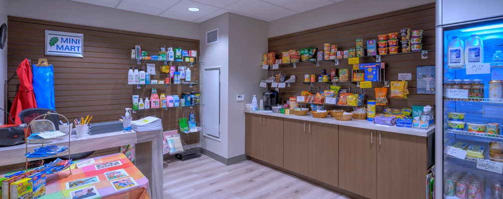 Photograph of interior room containing two tables with stationary items, shelves with products in bottles of various sizes, cabinets with nonperishable items on the counter, and a refrigerator stocked with perishable food.