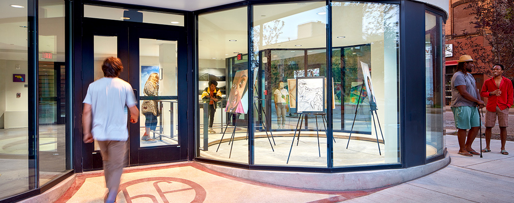 Photograph of a building entrance, with gallery space and art work behind plate glass windows. 
