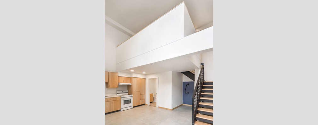 Interior photograph of a loft with stairs on the right, a kitchen with a stove and wooden cabinets on the left side, and a doorway to a bathroom in the background.