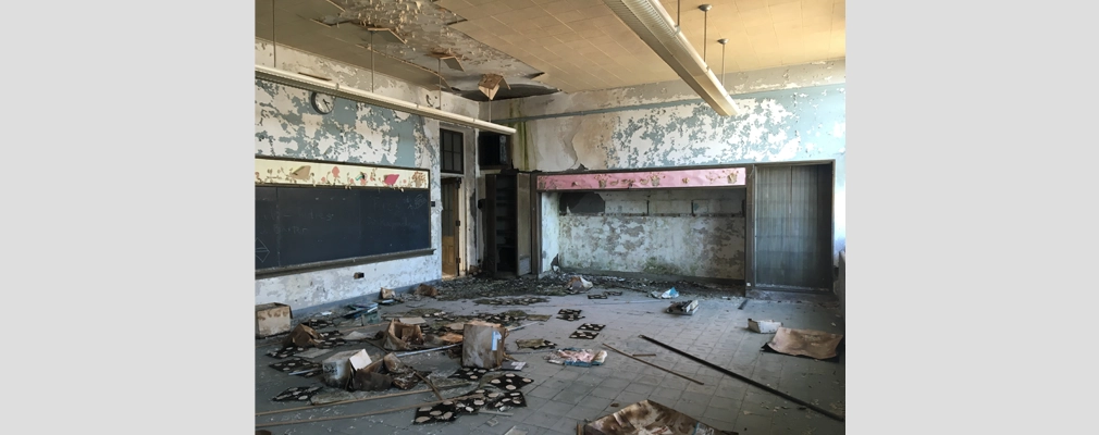 An abandoned, deteriorated classroom with tile flooring, damaged concrete walls, and trash scattered across the floor.