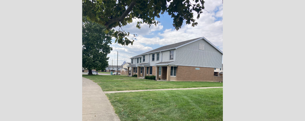 Photograph of the front and side facades of a row of two-story townhouses, taken from across a lawn.