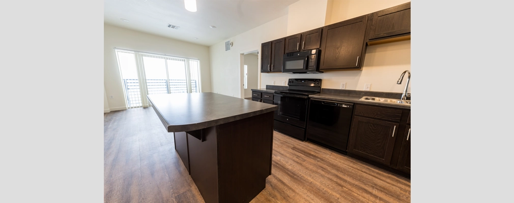 A kitchen with cabinets, appliances, and an island that opens into a living area with sliding doors to a balcony.