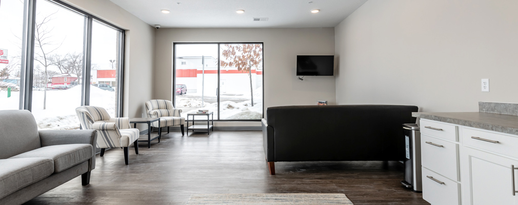 A large room with cabinets and a television area along one wall and an upholstered couch and two chairs arranged along another wall with large windows.