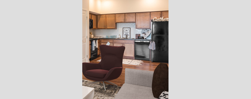 Photograph of a living room with a coffee table, chair, and sofa and a kitchen in the background.