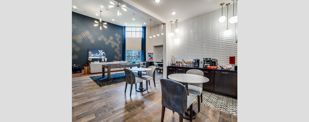 Community room with two tables with two chairs each in a kitchenette area with a lounge area in the background that includes a pool table, couch, and a television.