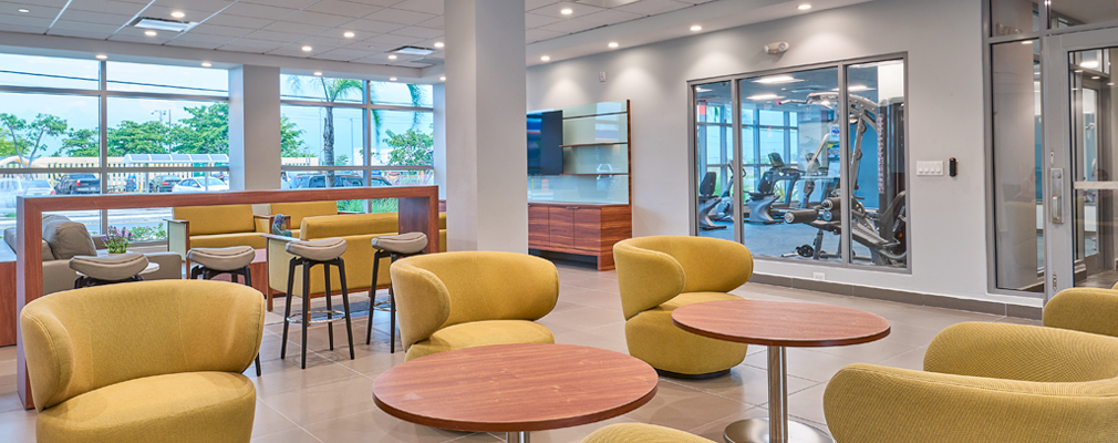 Photograph of upholstered chairs and tables in a spacious room, with large windows in two walls, one connecting to an exterior pool area and the other exposing a room with gym equipment. 