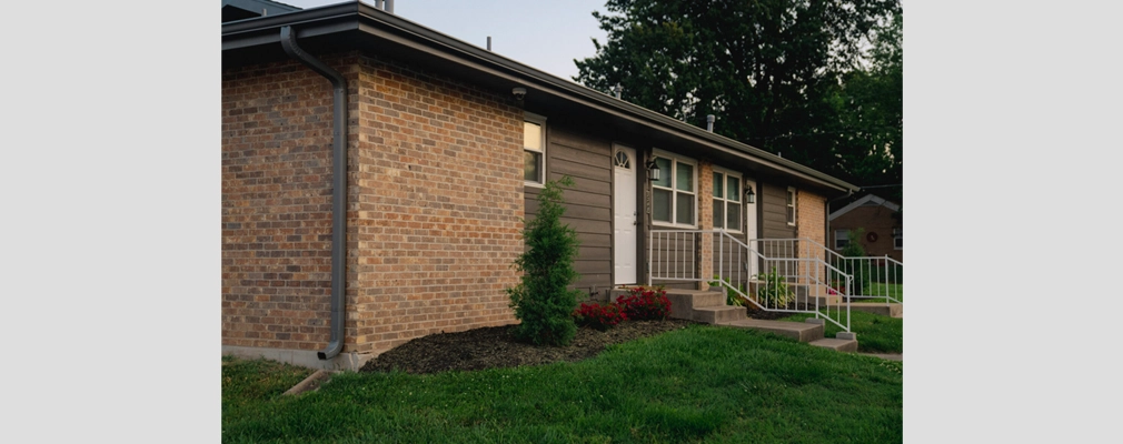 A photograph of a single-story, brick-and-siding residential building with exterior lighting, a landscaped front yard with flowers and shrubs, and a concrete walkway with metal railings leading up to the entrance.