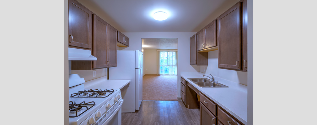 Photograph of interior view of kitchen with stove, sink, counter space, and upper and lower cabinets in the foreground and an unfurnished room and window in the background.