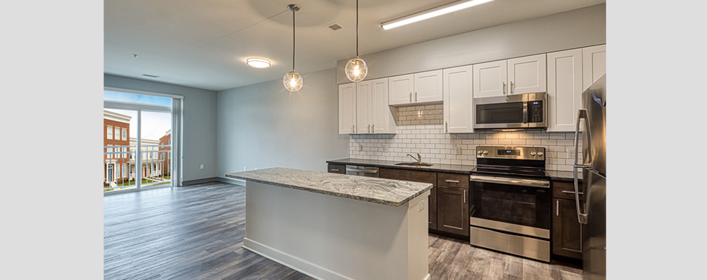 Photograph of a kitchen, with an island and upper and lower cabinets, and an unfurnished living area. 