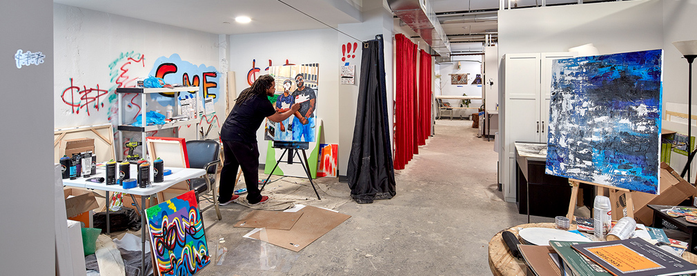 Photograph of an artist painting at an easel in a studio. 