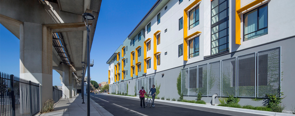 Photograph of two people walking on a paved path between elevated rail tracks on the left and a four-story multifamily building on the right, with screens covering wall openings for the first-floor garage and sunshades for the upper floor apartment windows.