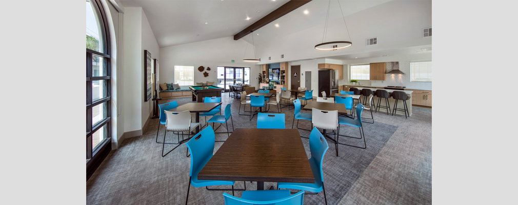 Photograph of an interior room with six sets of tables and chairs in the foreground and a kitchenette and living room in the background.